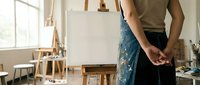 Female artist wearing paint-splattered apron holds brushes in bright studio workspace with easel and art supplies for creative painting projects.