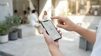 Person using smartphone with GPS navigation app in modern urban plaza with blurred people walking in background during sunny day.