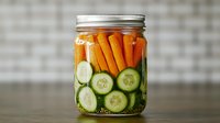 Fresh cucumber slices and baby carrots in glass mason jar with brine, arranged for pickling and fermentation. Healthy homemade preserves on rustic background.