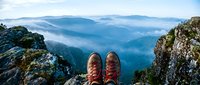 Hiker boots on rocky mountain summit overlooking misty blue valleys and distant peaks. Adventure travel and outdoor exploration concept for hiking gear.