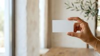 Woman with medium skin tone holding blank white business card in modern bright interior with natural light and potted plant, suitable for corporate branding and professional networking materials.
