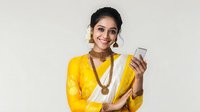 Young South Asian woman in bright yellow traditional sari and gold jewelry holding smartphone, smiling warmly against white background. Suitable for cultural and technology themes.