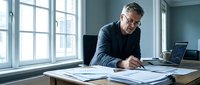 Mature male professional with glasses reviewing architectural blueprints and documents at wooden desk near bright windows in modern office space.