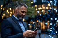 Mature bearded businessman in navy suit checking smartphone outdoors at dusk with festive bokeh lights creating atmospheric evening ambiance.
