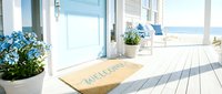 Coastal beach house entrance with welcome mat, blue door, white porch, ocean view, and blue flowers creating a serene summer retreat.