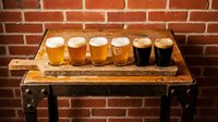 Beer flight tasting set with six glasses displaying variety of craft brews from light to dark on rustic wooden board against brick wall background for brewery, pub, or beverage industry use.
