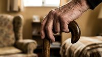 Elderly male hand with wrinkles and age spots gripping wooden cane handle indoors, representing senior mobility, aging, healthcare, and assisted living support for older adults.