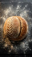 Artisan sourdough loaf with decorative scoring on dark floured surface, ready for bakery, culinary, or food photography projects.
