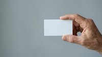 Senior male hand holding blank white business card against neutral gray background for corporate branding and professional networking purposes.