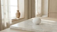Minimalist white marble table with ceramic egg and vase in bright sunlit room with flowing curtains creating serene modern interior design atmosphere.