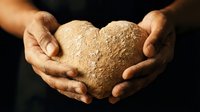 Hands gently holding heart-shaped whole grain bread loaf against dark background. Artisan bakery concept for healthy eating and homemade baking projects.