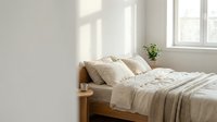 Wooden bed with beige linen bedding and pillows in bright, minimalist bedroom with natural sunlight from windows and potted plant on nightstand.