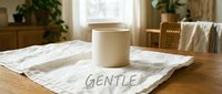 Cream-colored cylindrical candle jar displayed on white marble tile with "gentle" text, surrounded by potted plants and natural wooden furniture in bright interior space.