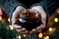 Woman hands holding glass jar with honey or amber liquid surrounded by warm golden Christmas lights and evergreen branches creating cozy holiday atmosphere.