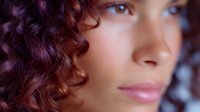 Close up portrait of young woman with purple curly hair and natural makeup against blurred background, soft lighting creates dreamy atmosphere.