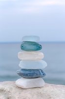 Stacked sea glass stones in white, blue, and green tones balanced on sandy beach with blurred ocean horizon, representing zen, balance, and coastal wellness themes.