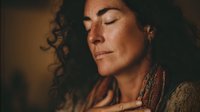 Middle-aged woman with curly dark hair in peaceful meditation pose, eyes closed, wearing bohemian style clothing against warm background.