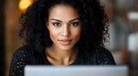 Young African American woman with natural curly hair smiling while working on laptop computer in cozy cafe setting with warm ambient lighting and soft bokeh background.