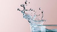 Dramatic water splash with droplets and ripples captured in high-speed photography against soft pink background, clear liquid motion in glass container.