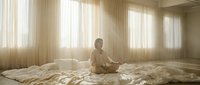 Young adult male with light skin tone in casual beige clothing sitting on unmade bed in bright, minimalist bedroom with sheer curtains and natural sunlight, conveying morning routine or contemplation.