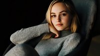 Young Caucasian woman with blonde hair wearing gray sweater reclining on dark chair, looking at camera with natural makeup and confident expression in moody lighting.