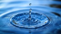 Crystal clear water drop splashing into blue water surface creating concentric ripple rings and crown splash formation with suspended droplets in macro photography.
