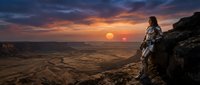 Futuristic female astronaut in spacesuit standing on rocky cliff overlooking alien desert landscape at sunset with dramatic orange and purple sky.