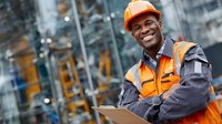 Happy African American construction worker in safety gear holding tablet at industrial site with modern building background.