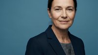 Middle-aged woman with dark hair, Caucasian ethnicity, wearing dark blazer over gray shirt against blue background. Professional portrait for business, corporate, and leadership concepts.