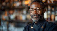Mature African American chef with glasses and gray beard smiling in restaurant with blurred bar background. Professional culinary portrait.