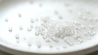 Transparent sugar crystals and granules scattered on white plate surface with shallow depth of field, suitable for food photography and culinary content.