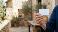 Man holding blank white business card outdoors in Mediterranean courtyard setting with potted plants and stone architecture for branding and corporate identity mockup.