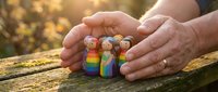 Adult hands cradling colorful wooden figurines representing diverse family members outdoors. Concept for inclusivity, acceptance, protection, and unity in relationships and communities.