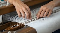 Hands working on traditional wooden loom weaving white threads creating textile fabric in artisan workshop craft process.