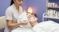 Professional spa therapist applying facial mask treatment to relaxed female client in modern wellness center with soft ambient lighting and skincare products.