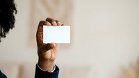 Male hand with dark skin tone holding blank white business card or paper against neutral background for branding and corporate messaging.