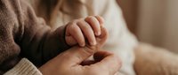 Tender moment of adult hands gently holding baby's tiny fingers in warm natural light. Family bonding, newborn care, parenting love and protection concept.