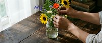 Person with medium skin tone arranging fresh sunflowers and mixed blooms in glass vase on wooden table by window, florist or home decor styling.