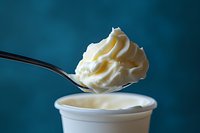 Creamy whipped topping on silver spoon hovering over white container against dark teal background, showcasing light, airy texture and smooth consistency.