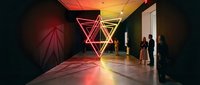 Modern art gallery interior with geometric neon light installation featuring triangular shapes in red and yellow colors creating ambient atmosphere.