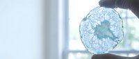 Blue agate slice with intricate crystalline patterns held against bright window light showcasing natural mineral formations and translucent gemstone texture.