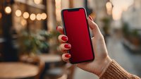 Female hand with red manicure holding smartphone with blank red screen in cozy cafe atmosphere with bokeh lights.