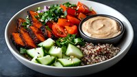 Fresh buddha bowl with grilled carrots, quinoa, cucumber, cherry tomatoes, and creamy hummus dip served in white ceramic bowl on dark background.