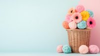 Pastel yarn balls and gerbera daisies in wicker basket against dual-toned pink and mint background, creating soft crafting composition for knitting and crochet projects.
