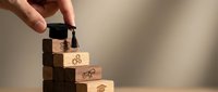 Hand placing graduation cap on wooden block staircase with education icons, symbolizing academic achievement and career growth steps.