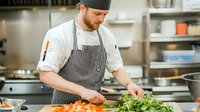Caucasian male chef in striped apron slicing fresh carrots on wooden cutting board in professional restaurant kitchen, preparing ingredients for cooking.