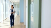 Professional Asian female doctor walking through modern hospital corridor with tablet, wearing white coat and stethoscope in bright medical facility.