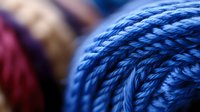 Extreme close up of vibrant blue yarn ball texture showing detailed wool fibers and twisted threads pattern against blurred burgundy background, suitable for crafting themes.