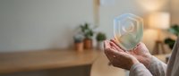 Close-up of mature hands holding glowing holographic shield symbol, representing data security, protection and digital safety in modern office environment with plants.