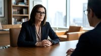 Professional businesswoman in glasses conducting interview or meeting with colleague in modern office setting with bookshelves and natural lighting.